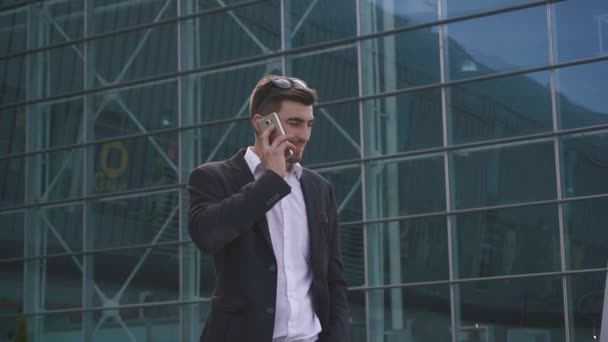 Confident young handsome bearded businessman talks by phone on the glass airport wall background - Video