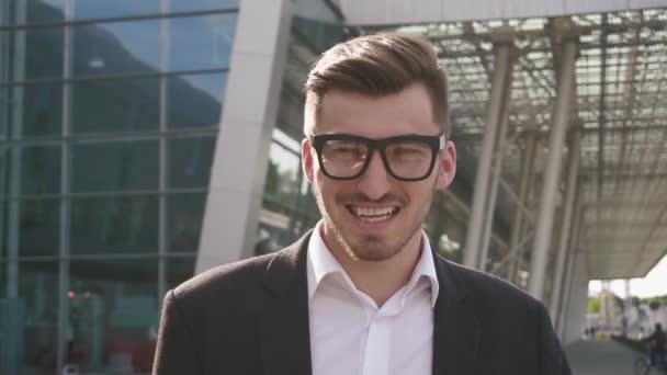 Close-up face of confident handsome smiling businessman adjusting glasses and staring at the camera. Outdoor airport background, business concept - Πλάνα, βίντεο