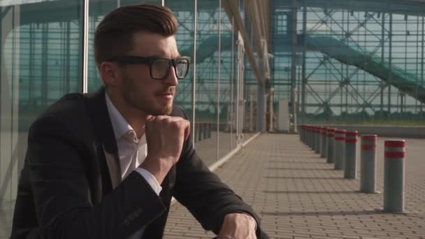 Close up portrait of an attractive businessman in glasses being worried and thoughtful while standing next to office buildings in a classic city square. - Felvétel, videó