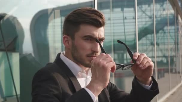 Close up portrait of an attractive businessman takes on glasses and being worried and thoughtful while standing next to office buildings in a classic city square. - Filmati, video