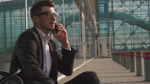 Urban styled businessman talking on smartphone while sitting at the airport and waiting for a flight. Casual young businessman wearing suit jacket and shoulder bag. - Felvétel, videó