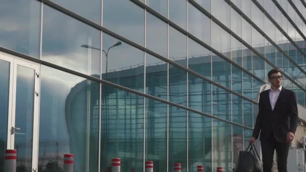 Attractive confident businessman in a modern stylish suit with luggage at the glass airport background - Filmagem, Vídeo