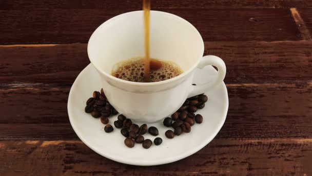eine Tasse Kaffee mit Kaffeebohnen auf Teller über braunen Holztisch gießen - Filmmaterial, Video