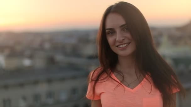 Seksowna dziewczyna z długimi włosami odwraca się i uśmiechnięta na zewnątrz kamery, niewyraźne miasto o zachodzie słońca na tle - Materiał filmowy, wideo