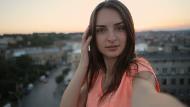 オレンジ色のTシャツで笑顔の女性は、夕方に素晴らしい街並みの景色を眺めながらテラスでカメラで自分撮り写真を撮り、クローズアップ - 映像、動画