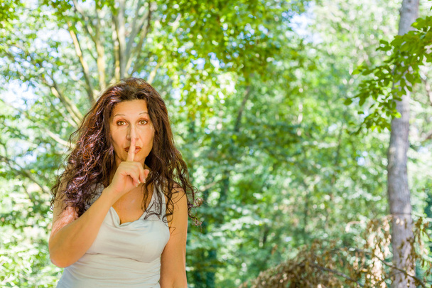 Καμπύλο αριστοκρατικό ώριμη γυναίκα θέσει δείκτη στο στόμα για τη σιωπή σε έναν κήπο - Φωτογραφία, εικόνα