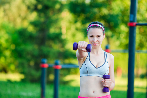 Beautiful young woman working out with weights outdoors. Active girl working out with small dumbbells in the park - 写真・画像
