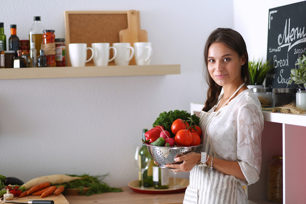Smiling young woman holding vegetables standing in kitchen - 写真・画像
