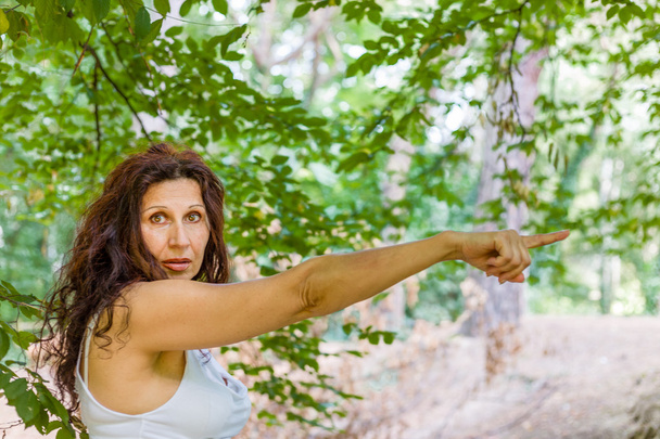 Kobieta w okresie menopauzy z zmarszczkami ubranych w białe i długie brązowe faliste włosy, wskazując palcem prawą rękę do jej lewej stronie patrząc w aparacie - Zdjęcie, obraz