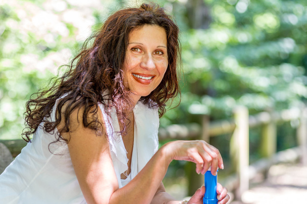 attractive woman in white smiles sitting on a park bench while opening the tube for soap bubbles - Photo, Image