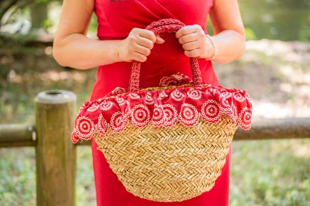 Während weibliche Hände eine Tasche im Landhausstil aus Bast und rotem Tuch mit weißen Blumen in der Hand halten, wartet die Frau im roten Mantel nervös in einem Park. - Foto, Bild