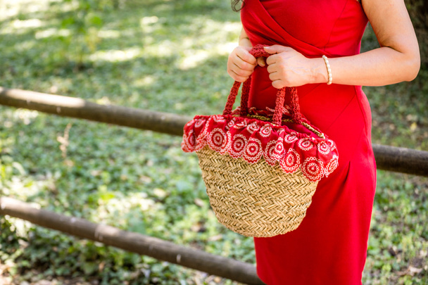 manos femeninas sosteniendo una bolsa en un estilo país hecho de rafia y tela roja con flores blancas, la mujer vestida con un vestido de vaina roja está esperando nerviosamente en un parque
 - Foto, Imagen