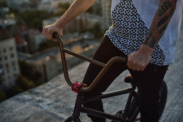 Молодий красивий хіпстер сидить на даху і позує на велосипеді на заході сонця
 - Фото, зображення