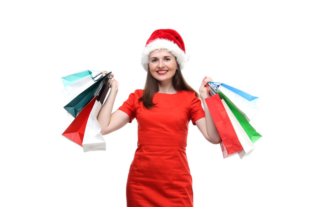 new year, cristmas, holidays concept - the portrait of young adult smiling woman wearing on red dress and Santa Claus hat  and holding colored shopping bags isolated on white background - Photo, Image