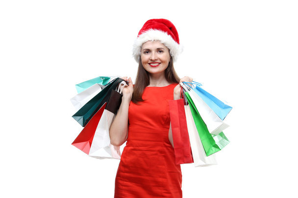 the portrait of young adult beautiful smiling woman wearing on red dress and Santa Claus hat holding colored shopping bags isolated on white background - Foto, Imagem