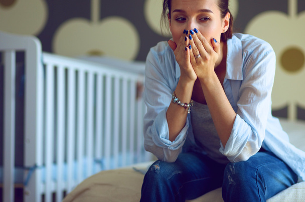 Young tired woman sitting on the bed near childrens cot. - 写真・画像