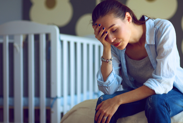 Young tired woman sitting on the bed near childrens cot. - Foto, Bild