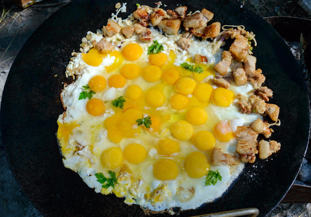 Τηγανιτά αυγά με τα κομμάτια του χοιρινού κρέατος για μια μεγάλη επίπεδη κατσαρόλα, μαγείρεμα σε εξωτερικούς χώρους - Φωτογραφία, εικόνα