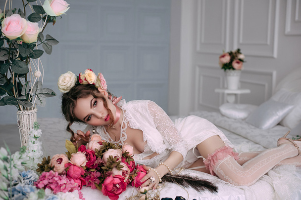 Beautiful young sexy woman sitting on white bed, wearing white lace dress, hair decorated with flowers. Perfect makeup. Beauty fashion. Eyelashes. Studio retouched shot.  - Photo, image