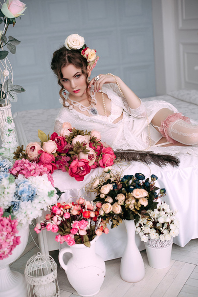 若いセクシーな美人白いベッドの上に座っている白いレースのドレス、花で飾られた髪を身に着けています。完璧なメイク。美容ファッション。まつげ。レタッチ スタジオ撮影.  - 写真・画像