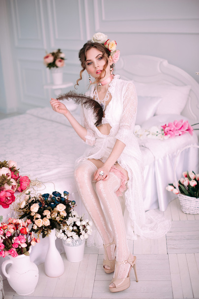 Όμορφη νεαρή σέξι γυναίκα κάθεται στο λευκό κρεβάτι, φορώντας λευκή δαντέλα φόρεμα, μαλλιά, διακοσμημένο με λουλούδια. Τέλειο μακιγιάζ. Ομορφιά μόδα. Βλεφαρίδες. Studio retouched βολή.  - Φωτογραφία, εικόνα