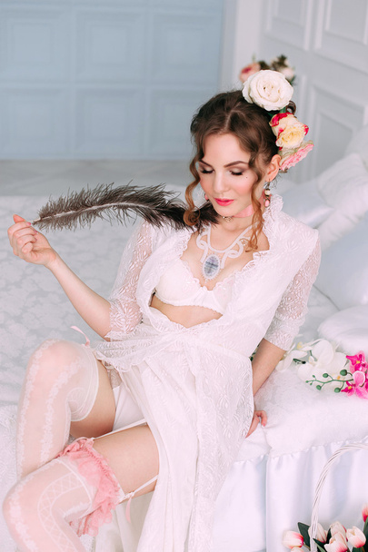 Szép fiatal szexi nő ül a fehér ágy visel fehér csipke ruha, haj, virágokkal díszített. Tökéletes smink. Szépség-divat. Szempillák. Stúdió retusált lövés.  - Fotó, kép