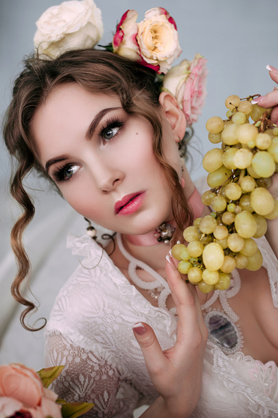 Красива молода сексуальна жінка сидить на білому ліжку і їсть виноград, одягнений в біле мереживне плаття, кімнату, прикрашену квітами. Ідеальний макіяж. Красива мода. Вії. Студійний ретушований знімок
.  - Фото, зображення