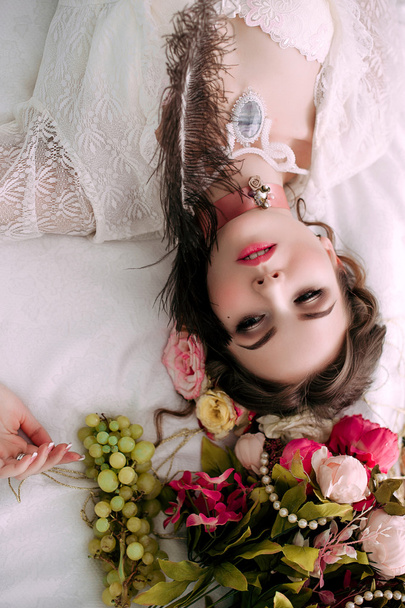 Hermosa mujer sexy joven sentado en la cama blanca y comer uvas, con vestido de encaje blanco, habitación decorada con flores. Maquillaje perfecto. Moda de belleza. Pestañas. Estudio retocado tiro
.  - Foto, Imagen