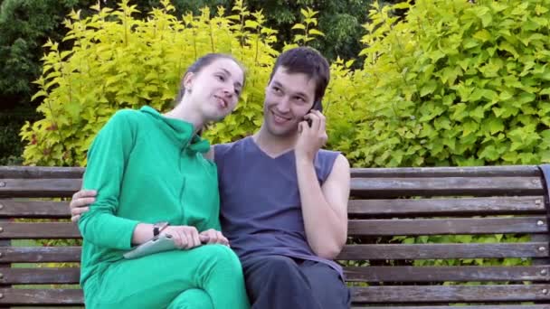 schönes junges Paar, das sich auf einer Parkbank ausruht. Sie umarmen sich und der Mann telefoniert. - Filmmaterial, Video