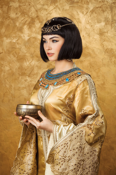 Beautiful woman like Egyptian Queen Cleopatra with cup on golden background - Photo, image
