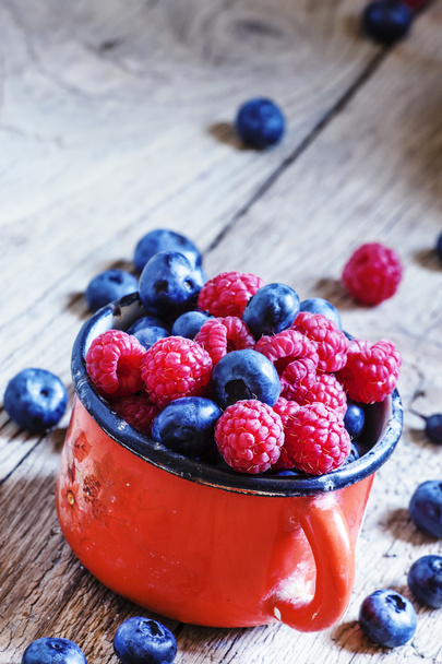 Fresh raspberries and blueberries in red enamel mug - Fotó, kép