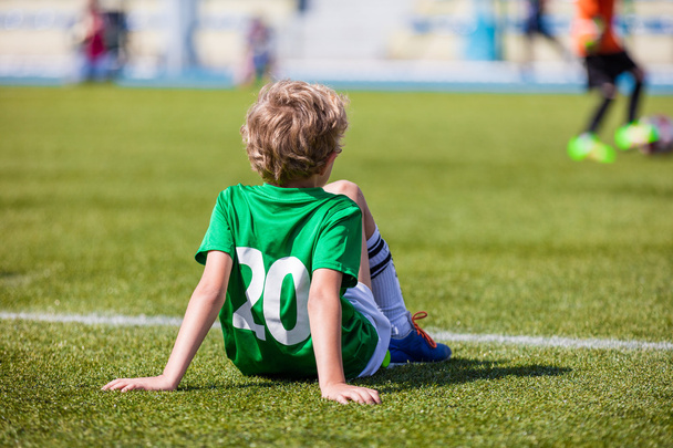 Nuorten jalkapallo, pieni jalkapalloilija. Nuori poika jalkapalloilija istuu jalkapallokenttä ja katsomassa koulun jalkapallo ottelu
 - Valokuva, kuva