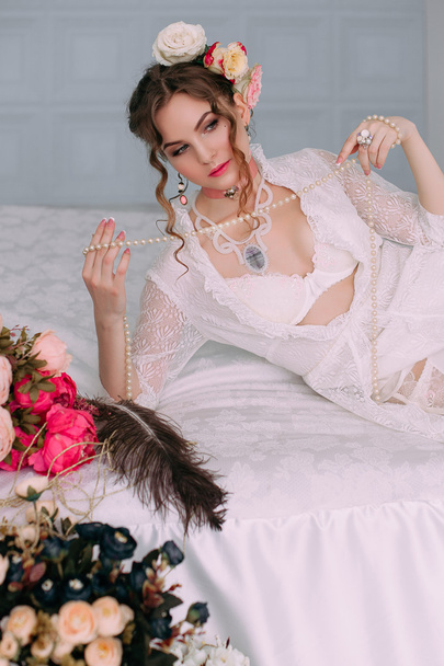 Szép fiatal szexi nő ül a fehér ágy visel fehér csipke ruha, haj, virágokkal díszített. Tökéletes smink. Szépség-divat. Szempillák. Stúdió retusált lövés.  - Fotó, kép