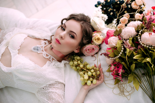 Krásná mladá sexy žena sedí na bílé posteli a jíst hrozny, na sobě bílé krajkové šaty, místnosti vyzdobené květinami. Perfektní make-up. Móda krása. Řasy. Studio retušované shot.  - Fotografie, Obrázek