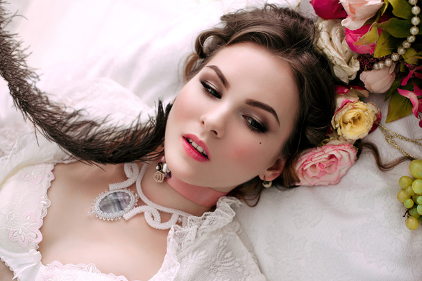 Krásná mladá sexy žena sedí na bílé posteli a jíst hrozny, na sobě bílé krajkové šaty, místnosti vyzdobené květinami. Perfektní make-up. Móda krása. Řasy. Studio retušované shot.  - Fotografie, Obrázek