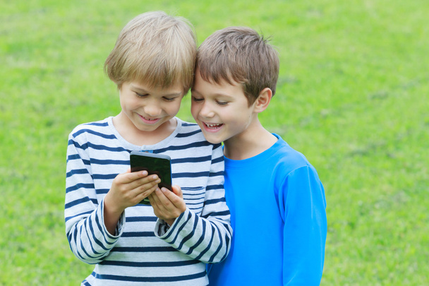 Children with mobile phone outdoor. Two boys smiling, looking to screen, playing games or using application. Technology education leisure people concept - Zdjęcie, obraz