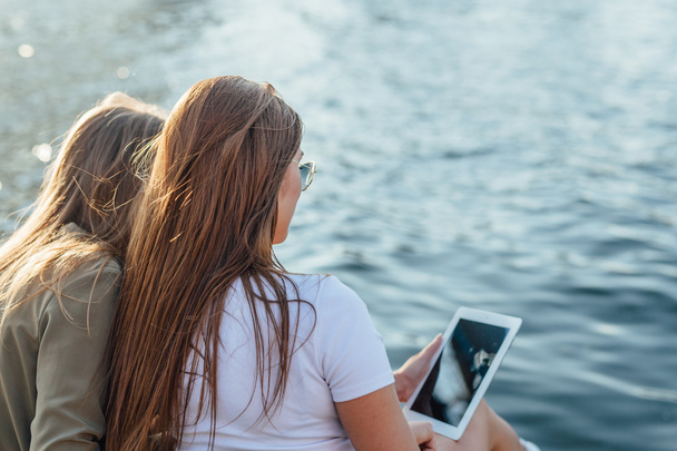 Two young girls sitting next to the river and using tablet pc. - Photo, image