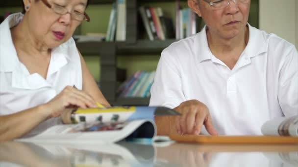 Asian senior couple reading books and magazines together in small library. Learning and study concept - Felvétel, videó