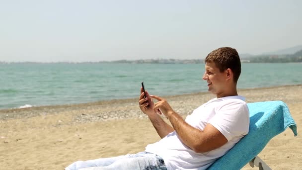 Homme sur Internet avec smartphone sur la plage. L'homme surfe sur Internet sur un fond de mer. L'homme se trouve sur une chaise longue avec un smartphone. Un homme se reposant sur la mer, regardant le téléphone
. - Séquence, vidéo