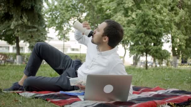 Joven ejecutivo trabajando en el parque en el portátil, mientras almorza
 - Metraje, vídeo