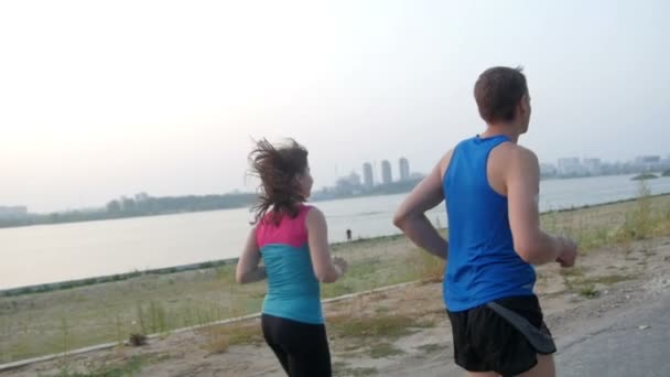 Couple of young adult athletes: woman and man running along promenade of river in modern city. Healthy lifestyle concept, slow-motion, rear view - Materiał filmowy, wideo