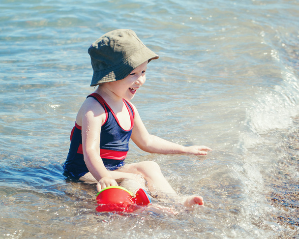 Portrait of cute adorable happy smiling toddler Caucasian girl with hat and watering pot toy on beach sitting in water waves having fun, emotional face expression, lifestyle summer mood - Photo, Image