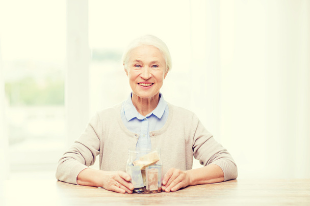 senior woman with money in glass jar at home - Foto, Imagem