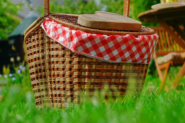 Primer plano de la cesta de picnic o cesto con paño a cuadros en el césped patio trasero. Corazón de madera en la cubierta de la cesta
 - Foto, Imagen