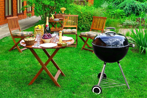 Καλοκαίρι BBQ οικογενειακή σκηνή πάρτι στον διακοσμητικούς κήπους στην πίσω αυλή. Συσκευή σχάρας ξυλάνθρακα, ξύλινες καρέκλες και τραπέζι με ορεκτικά και κρασί σαμπάνιας στο φρέσκο γκαζόν - Φωτογραφία, εικόνα