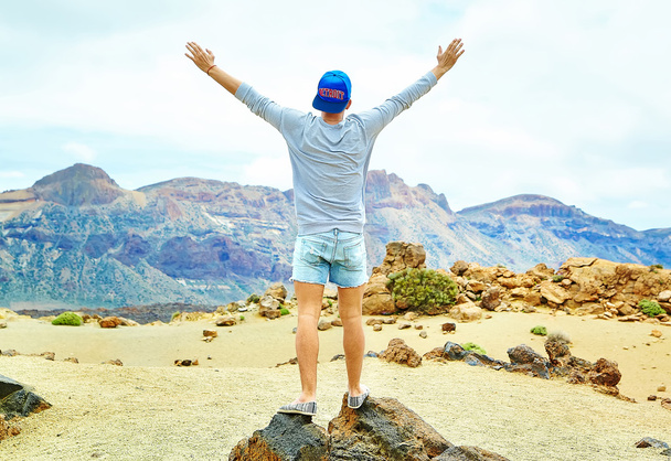 Happy stylish man in casual hipster clothes standing on the cliff of mountain with raised hands to the sun and celebrating success - Photo, image
