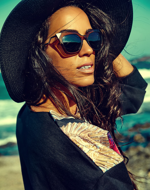 fashion stylish beautiful young smiling brunette woman model in summer hipster casual clothes posing behind blue ocean and sky background in hat and sunglassesfashion stylish beautiful young smiling brunette woman model in summer hipster casual cloth - Fotoğraf, Görsel