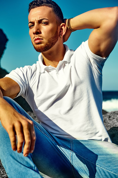Young stylish handsome man model in hipster summer clothes posing on beach behind sky and ocean - 写真・画像
