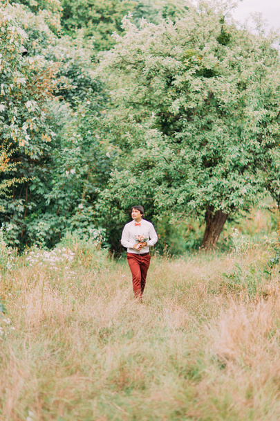 Элегантный стильный молодой жених в кирпичном цвете брюк с букетом цветов, гуляющий один в парке
 - Фото, изображение