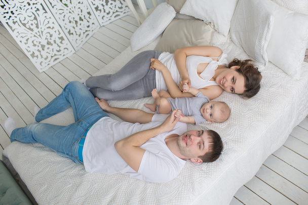 Ansicht von oben: Junge Familie liegt mit ihrem kleinen Baby auf einem Bett. Kleidung - weiße Hemden und Jeans - Foto, Bild